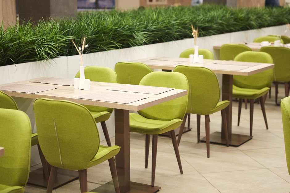 Interior view of a café seating area featuring a dark upholstered bench with a high back and slender armrests, positioned along a wall with large windows. In front of the bench are two round wooden t