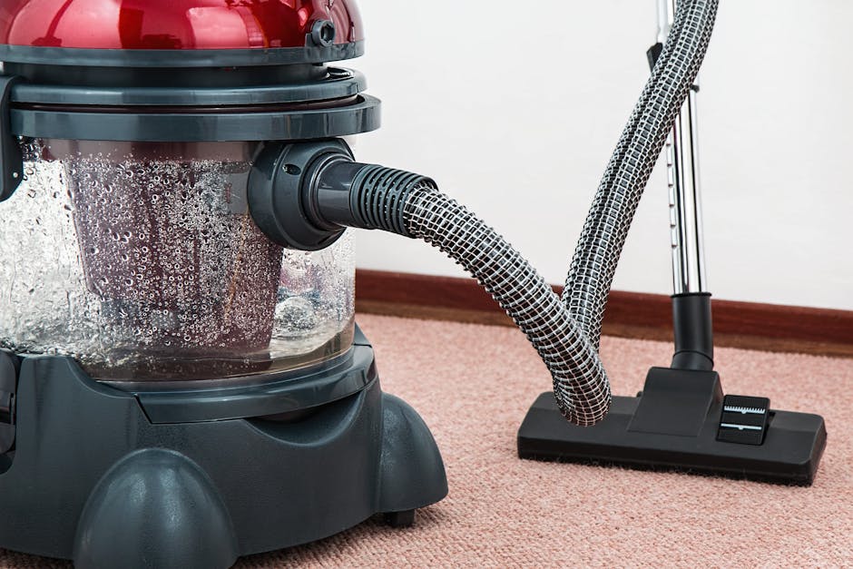 Close-up image of a vacuum cleaner with a transparent dust container filled with debris, situated on a pink carpet in a residential room. The vacuum's hose and wand are attached, with the wand resting on the carpet. The carpet appears clean, and the room features a white wall and a wooden skirting board. The vacuum cleaner is branded 'Carpet Cleaners N4' and is positioned to demonstrate surface cleaning and deep cleaning processes for domestic environments, showcasing equipment used for routine sanitisation and maintenance of home carpets.