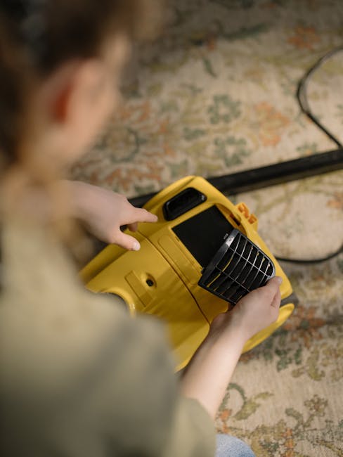 A person holding a yellow portable commercial carpet extractor with a black vented attachment, preparing for deep cleaning on a patterned carpet with floral motifs in a residential or commercial space. The extractor is positioned on the floor, and a black power cord extends from it, indicating an electrical connection. The scene is well-lit, highlighting the cleanliness of the surface. The background features a textured wall, and the person's hands are engaged with the equipment, emphasizing the focus on surface cleaning and maintenance. The image aligns with professional cleaning practices offered by Carpet Cleaners N4, specializing in sanitisation and deep cleaning for properties near Finsbury Park Station, N4.