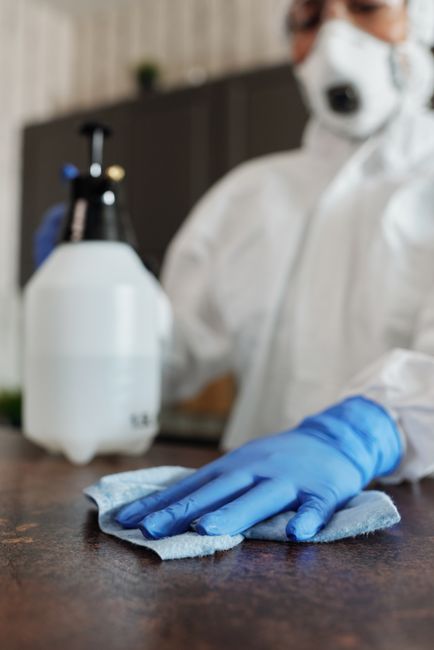 A professional cleaner at Carpet Cleaners N4 is performing surface cleaning and sanitisation in an indoor environment, wearing full protective gear including a white coverall, mask, goggles, and glove