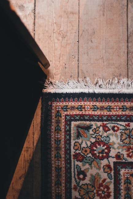 A close-up view of a corner in a room featuring a patterned rug with intricate floral designs in shades of red, blue, yellow, and cream, partially covering a wooden floor with a natural finish. The rug's edge has a fringe, and the wooden floor shows visible grain and plank lines. The scene is illuminated with soft, natural lighting, highlighting the cleanliness and well-maintained condition of the surfaces. The bottom left corner of a piece of dark furniture is visible, suggesting a domestic setting. This image exemplifies surface cleaning and maintenance, consistent with the services provided by Carpet Cleaners N4 for domestic cleaning and rug sanitisation on Stroud Green Road in Finsbury Park, N4.