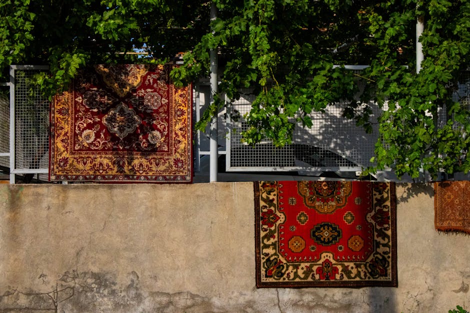 An outdoor balcony area featuring three intricately patterned oriental rugs with red, gold, and cream tones arranged on a concrete floor. The balcony is bordered by metal railings with a grid design, partially covered by lush green foliage from a vine sprawling across the top. Bright sunlight casts shadows on the rugs and the textured, slightly weathered concrete surface, highlighting the cleanliness and vibrant colors of the rugs, which appear to be well-maintained. The scene suggests a space for relaxation or outdoor sitting, with natural light emphasizing the detail and condition of the textiles, and the greenery adding a touch of natural freshness. Carpet Cleaners N4 specializes in surface cleaning and deep cleaning services, including sanitisation, to maintain hygiene and aesthetic appeal of domestic and commercial spaces.